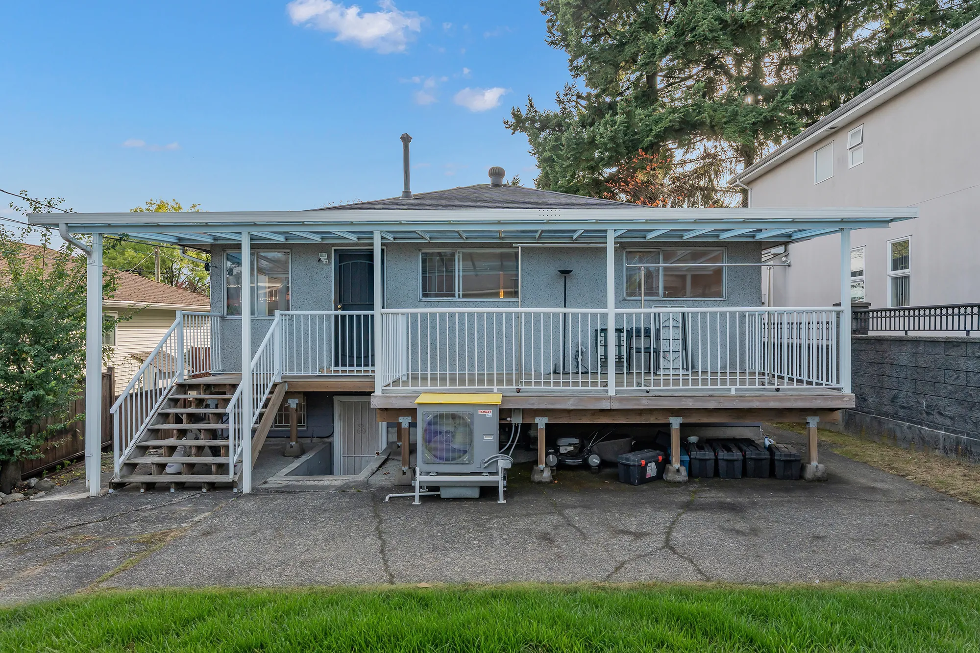 The back of the house: a covered deck across the main floor with white railings and stairs down to the yard, a heat pump unit under the deck, a concrete yard