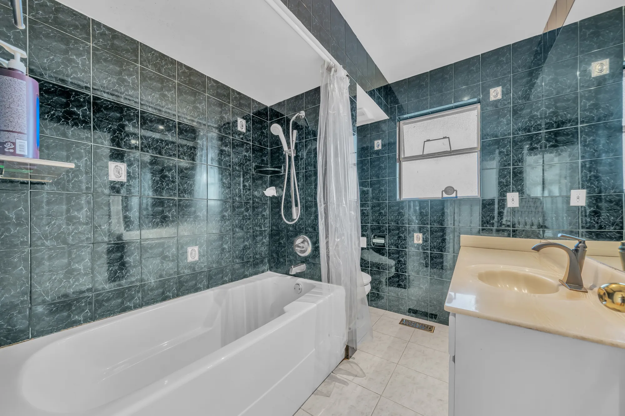 The main bathroom: a tub with a shower, dark green marble-look tile walls, a vanity with a cream top, a window