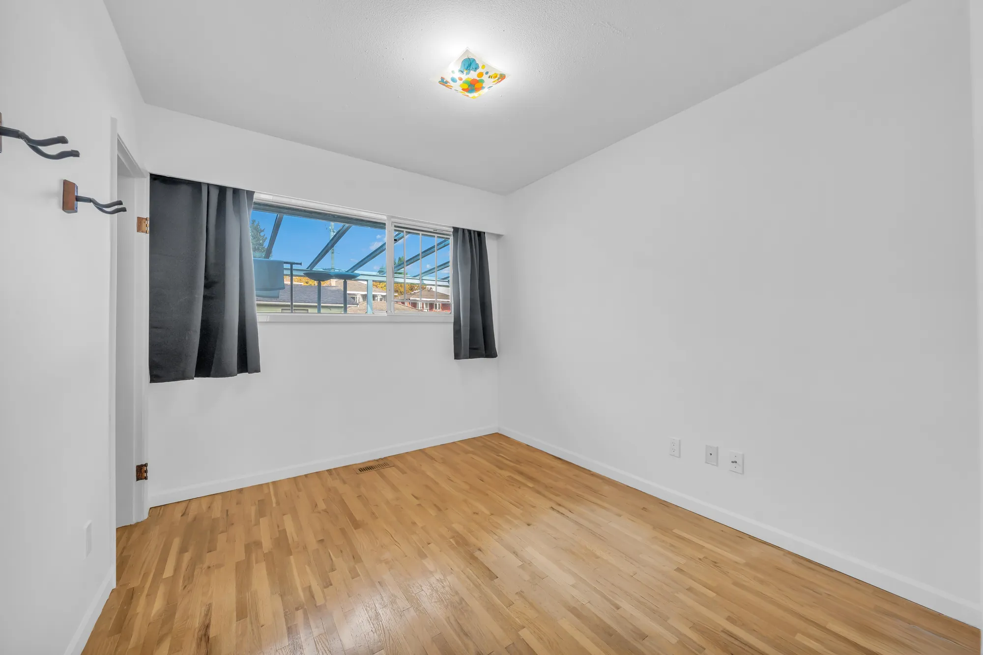 A bedroom, empty: a wood floor, a window with dark curtains onto the clear roof of the deck, coat hooks by the door