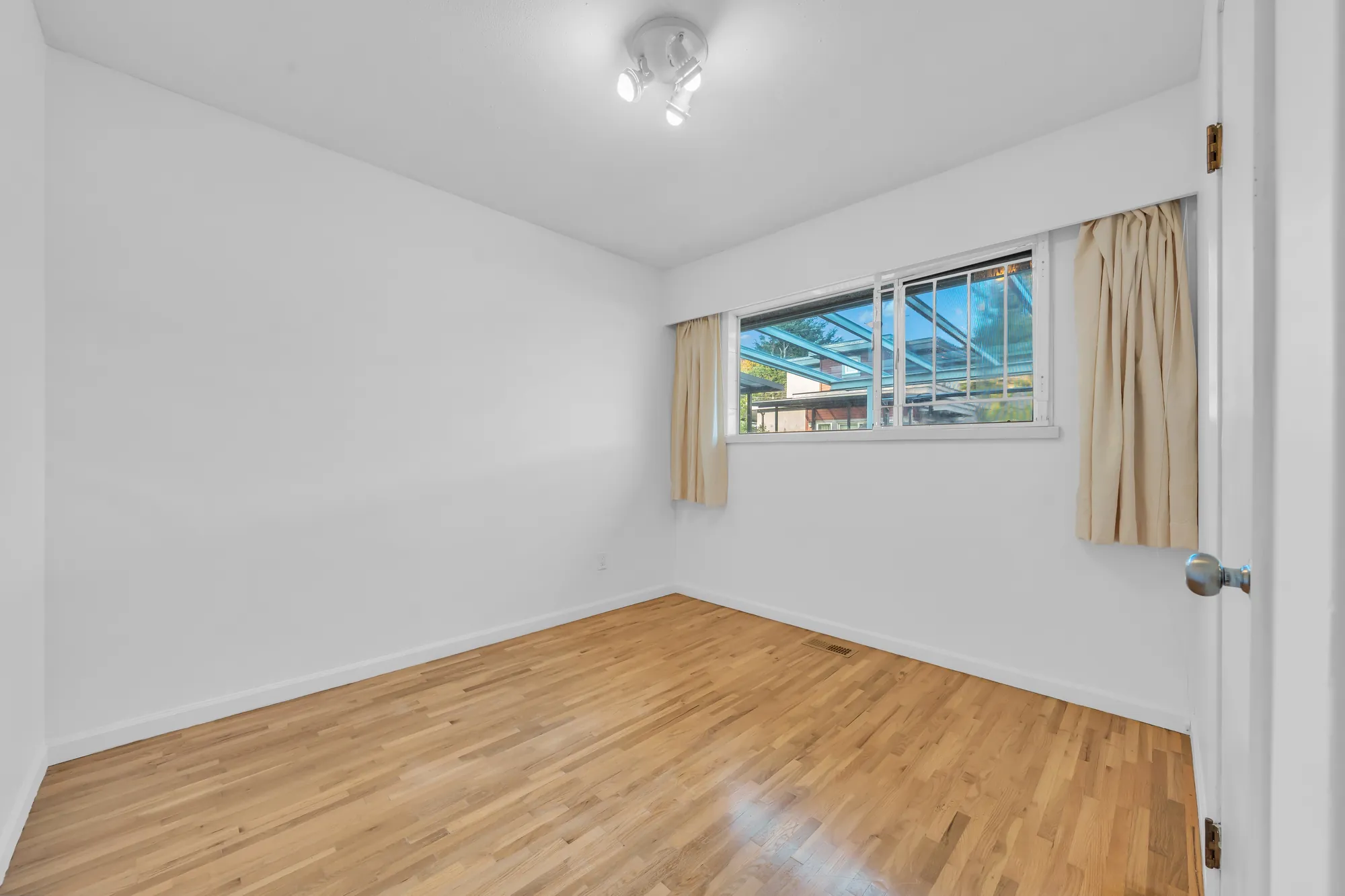 The other back bedroom, empty: a wood floor, a window with beige curtains onto the clear roof of the deck, a track light