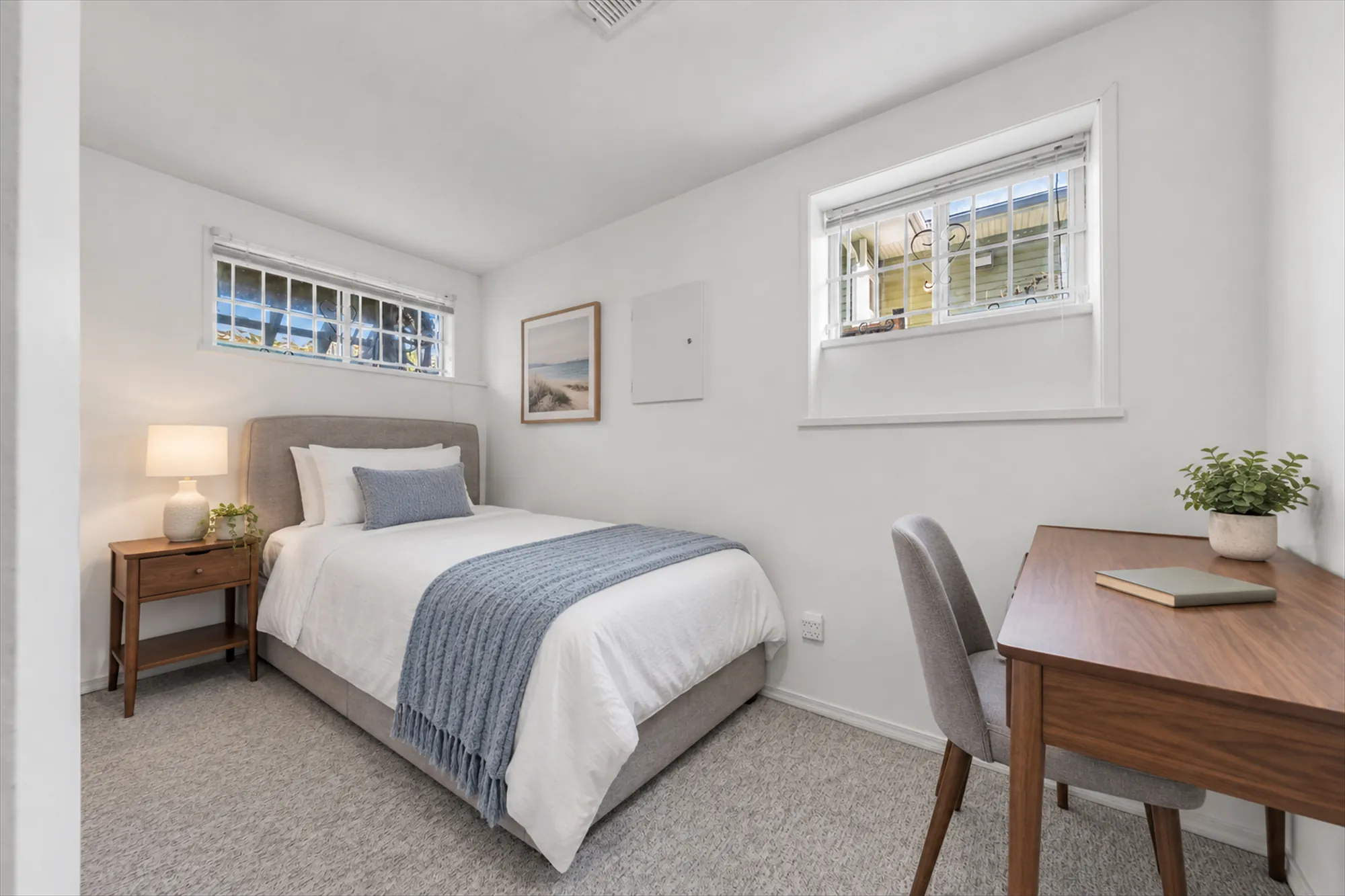A lower bedroom with two windows, virtually staged: a bed with a blue throw, a night table, a desk and chair, a plant