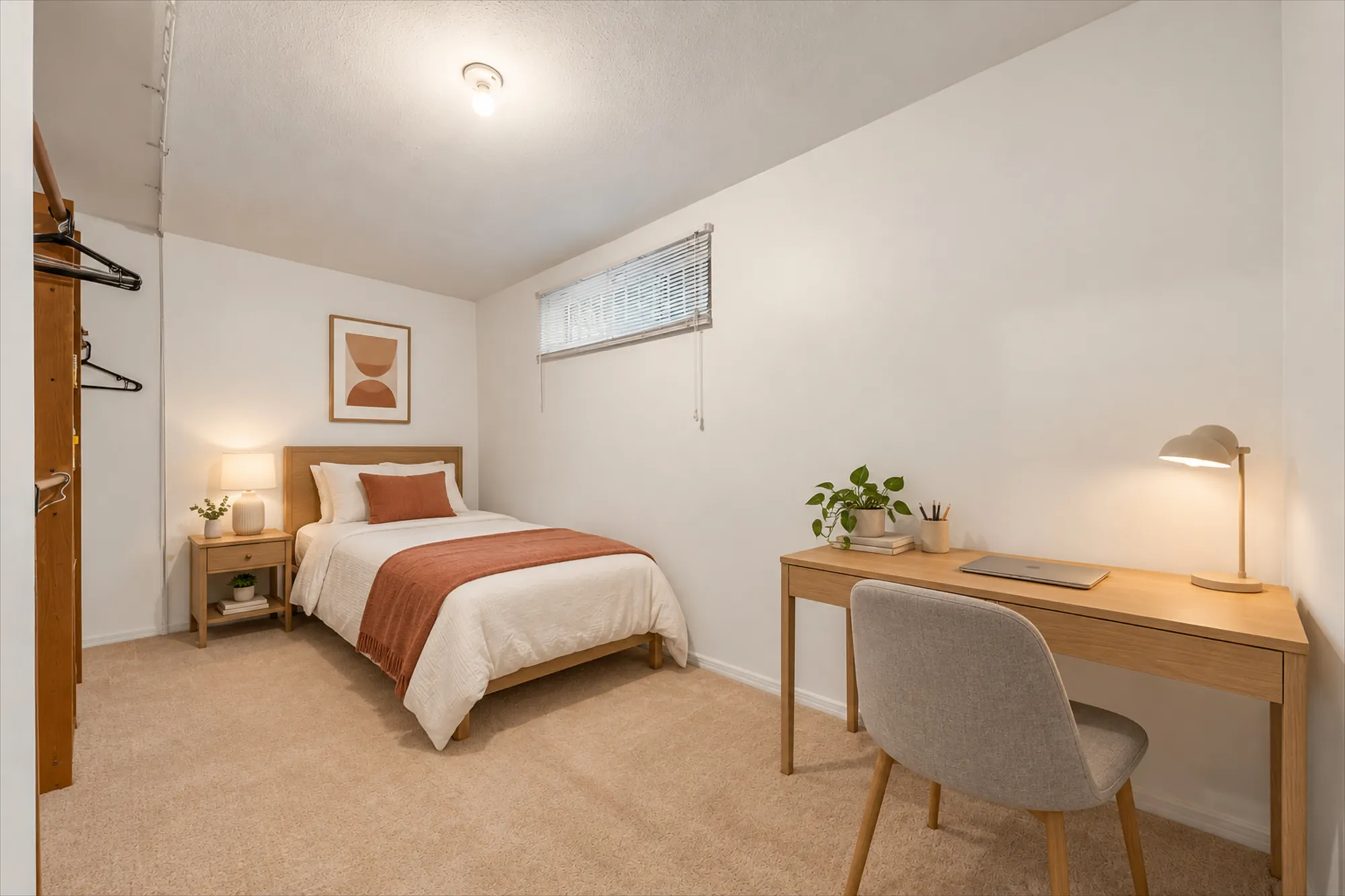 A lower bedroom, virtually staged: a bed with a rust throw, a night table, a desk with a lamp and a chair, an open closet with rails at the left