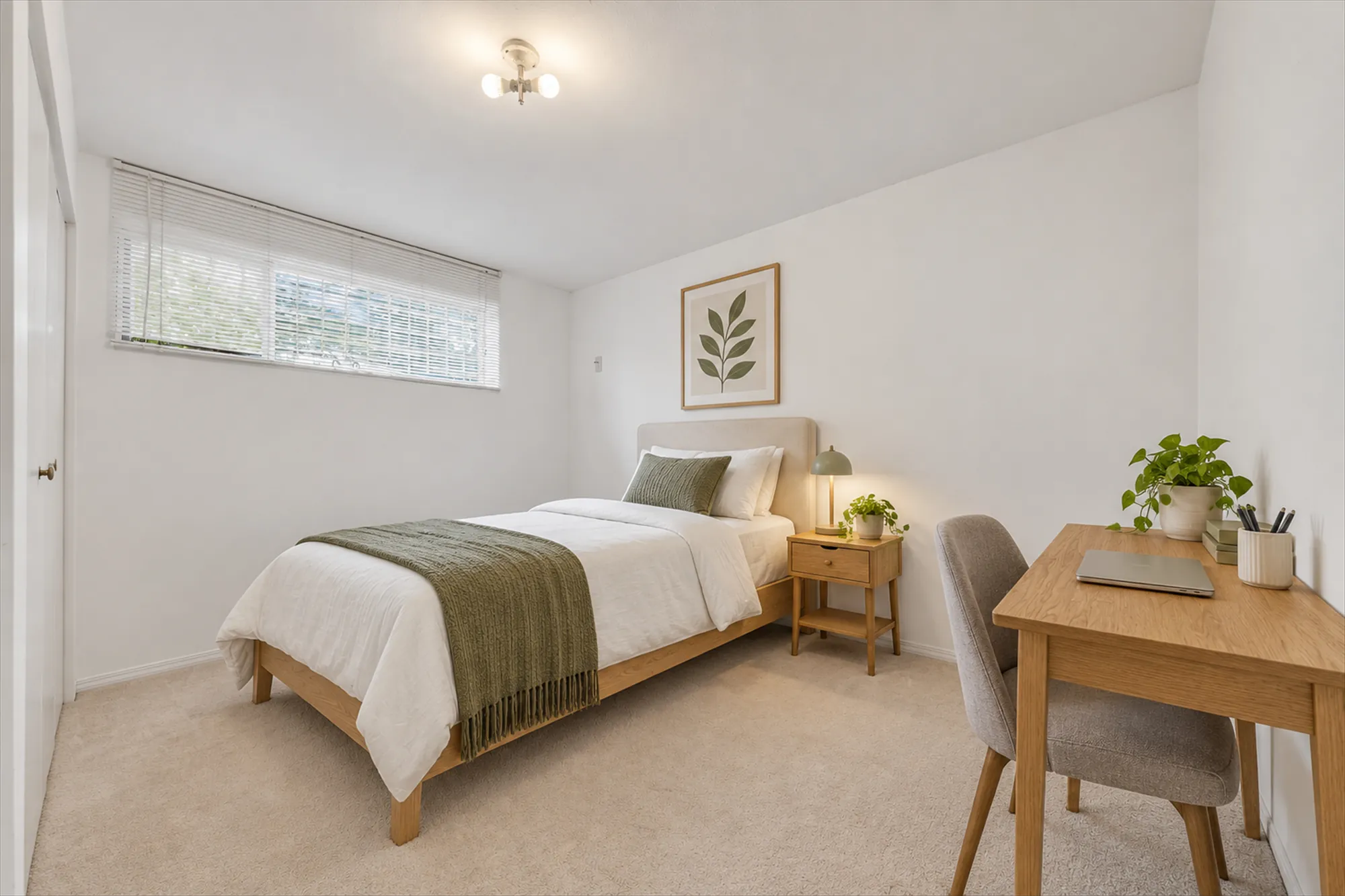 A lower bedroom, virtually staged: a single bed with a green throw, a night table, a desk and chair, a plant, a botanical print