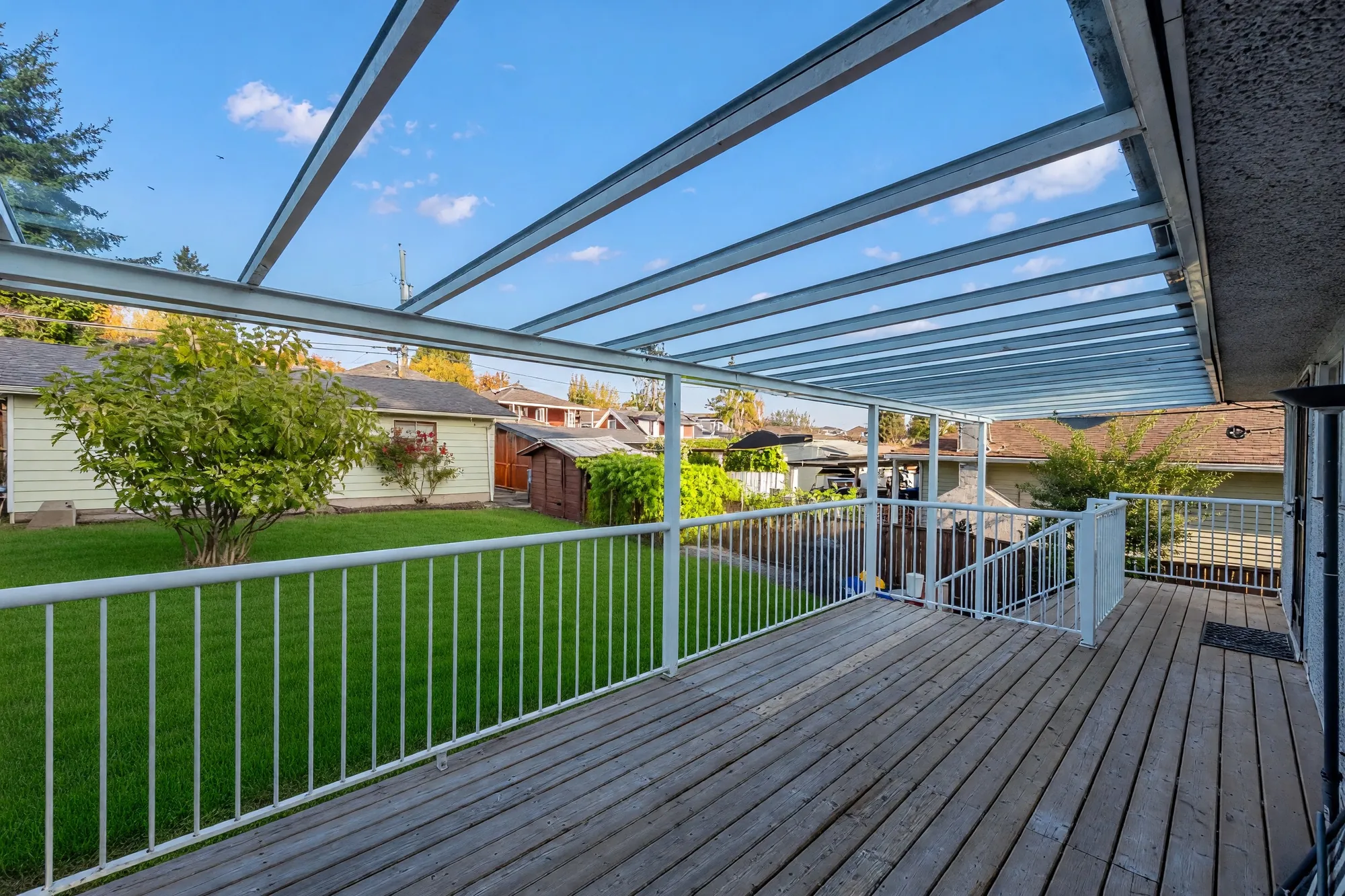 The covered deck: a clear roof on white beams, white railings, wood boards, the back lawn and the garage beyond