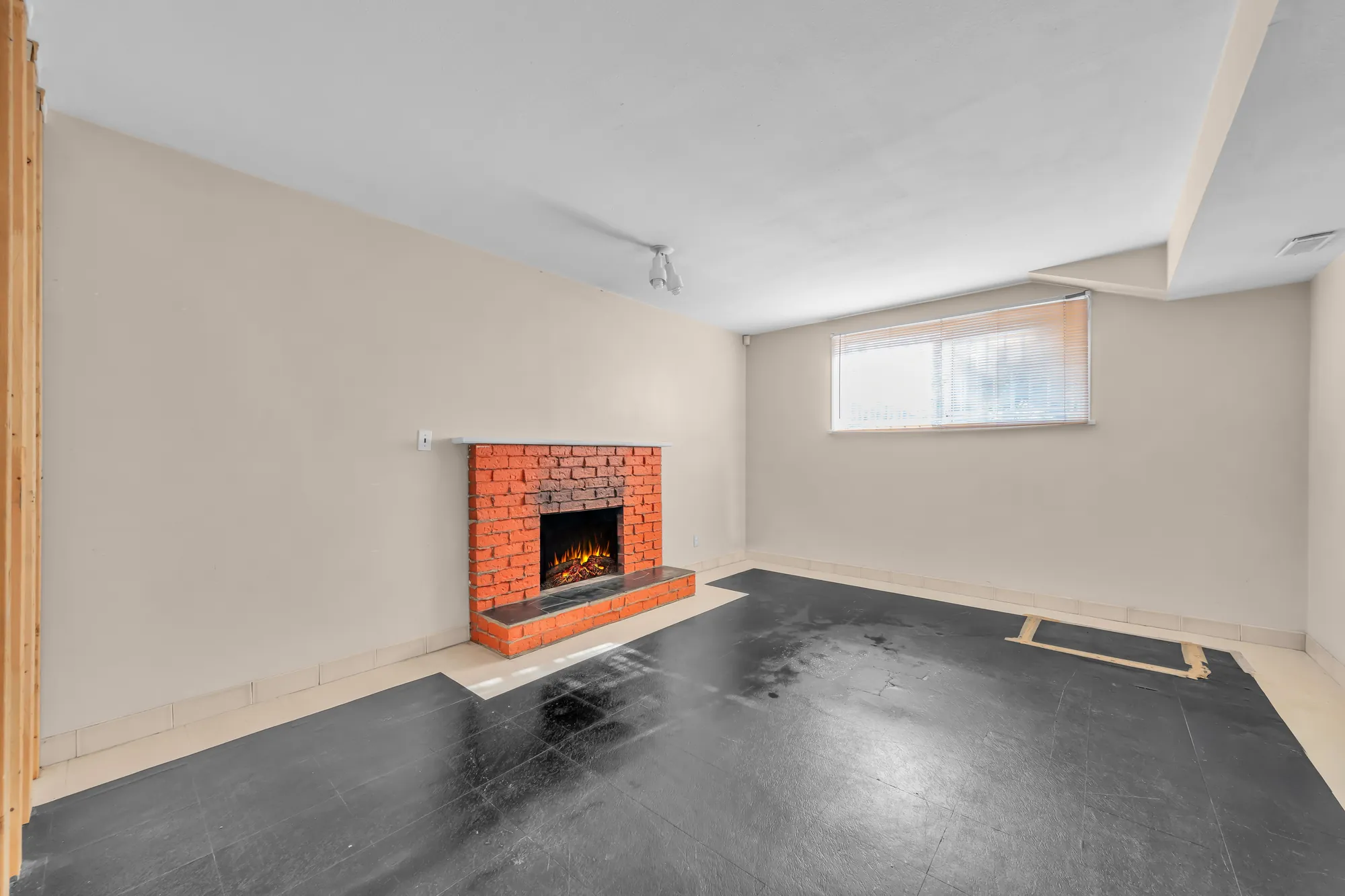 The lower family room, empty: a brick fireplace with an insert, a dark vinyl floor, a window with blinds