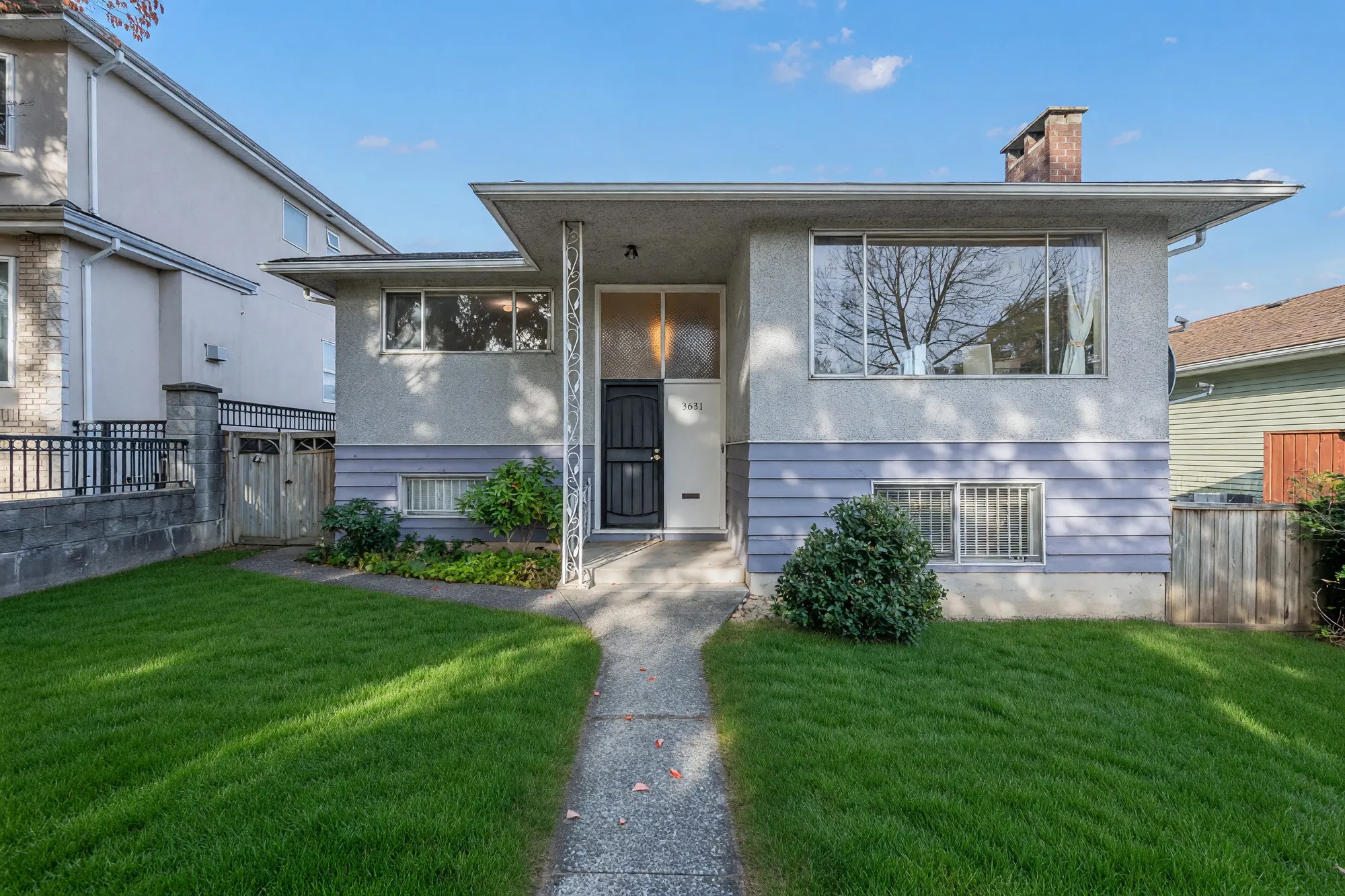 The front of the house from the walk: a stucco upper floor with a wide picture window over a lower floor in lavender-grey siding, a black security door beside the number 3631, a concrete path between two lawns, a brick chimney