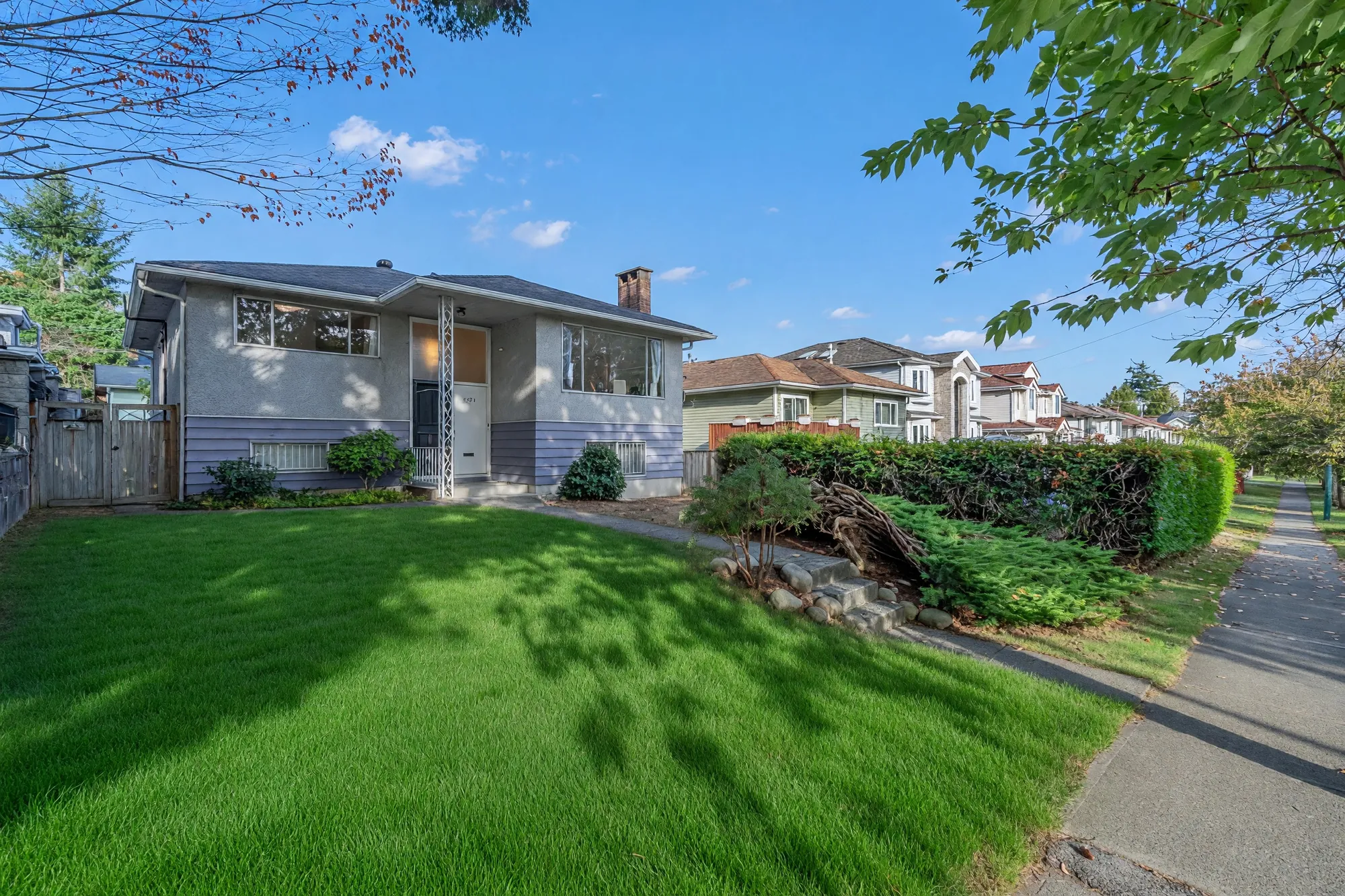 The house from the corner of the lot across the front lawn, a clipped hedge along the side, the sidewalk at the right, neighbouring houses beyond