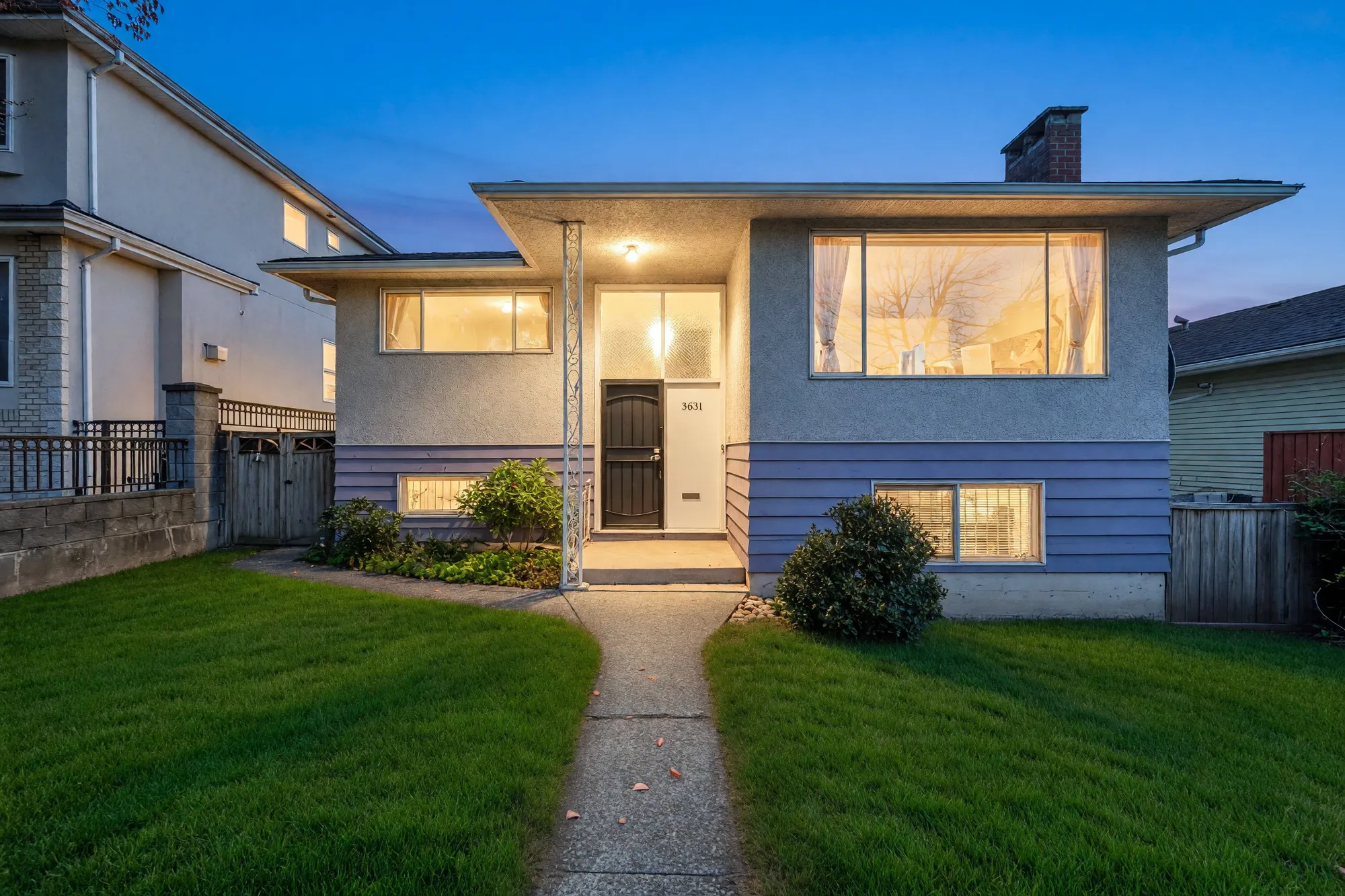The front of 3631 Wellington Avenue at dusk: a stucco upper floor with a lit picture window over a lower floor in lavender-grey siding, a light over the front door, a concrete path between two lawns