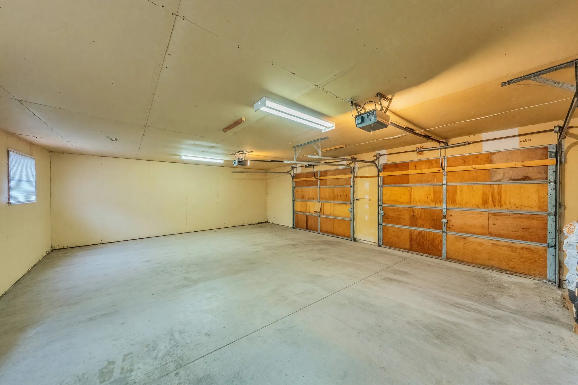 Inside the double garage, empty: a concrete floor, wood-panelled overhead doors with an opener, a fluorescent light, a window