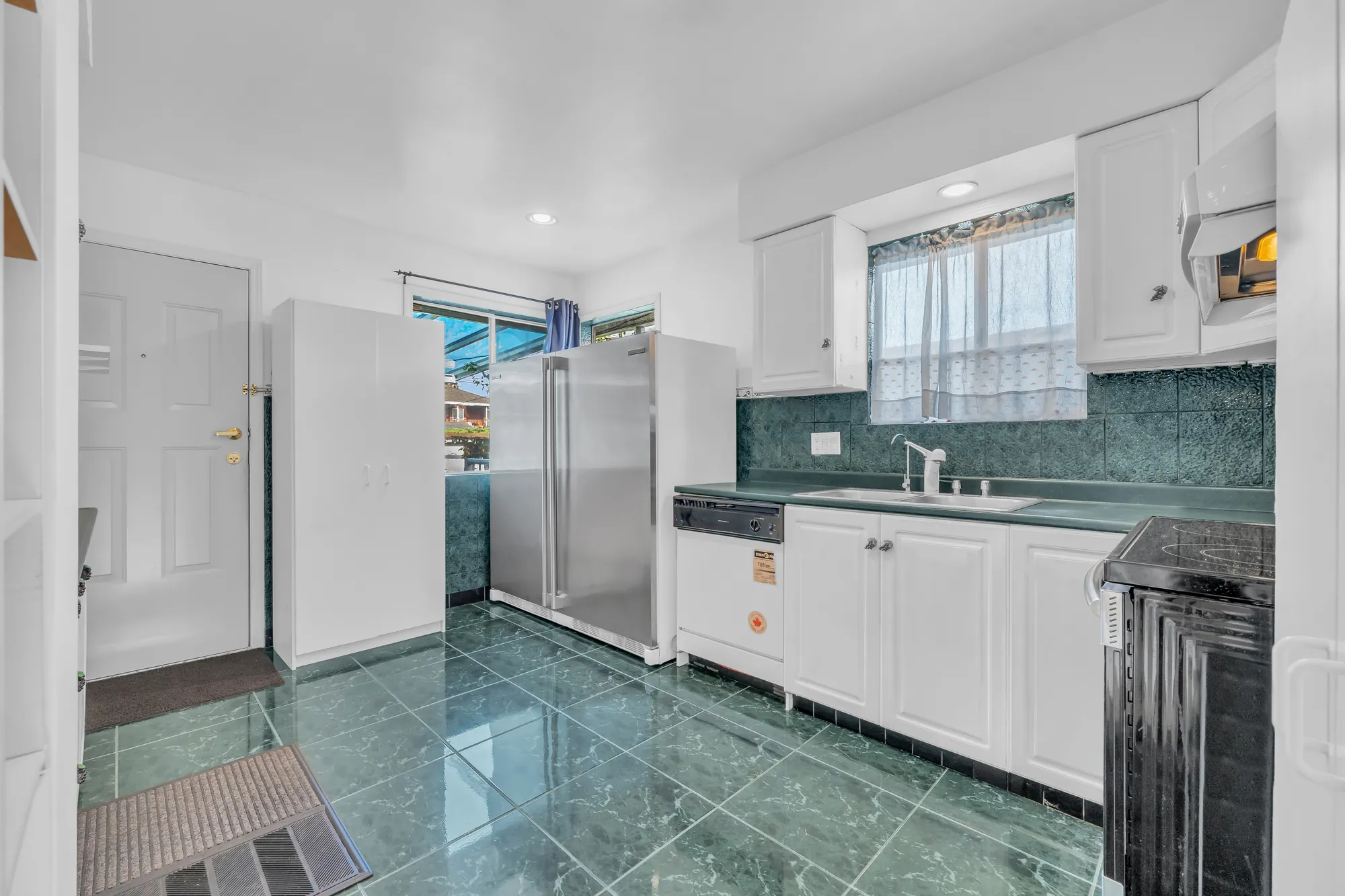 The kitchen toward the back door: a tall white cabinet, the refrigerator, a dishwasher and the sink under a window with lace curtains, a window onto the covered deck