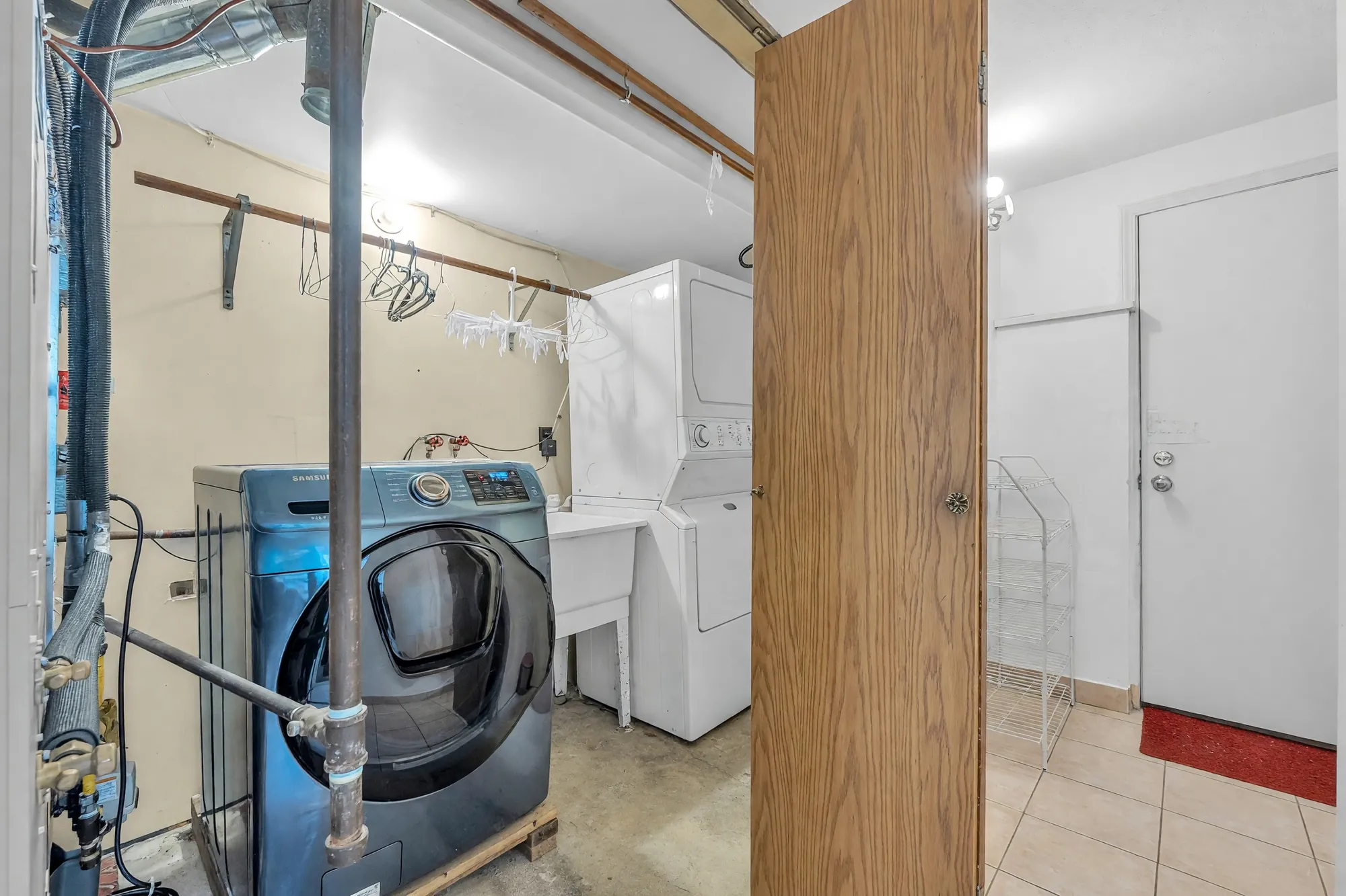 The utility room: a front-loading washer, a stacked washer and dryer, a laundry sink, exposed pipes, a wood door