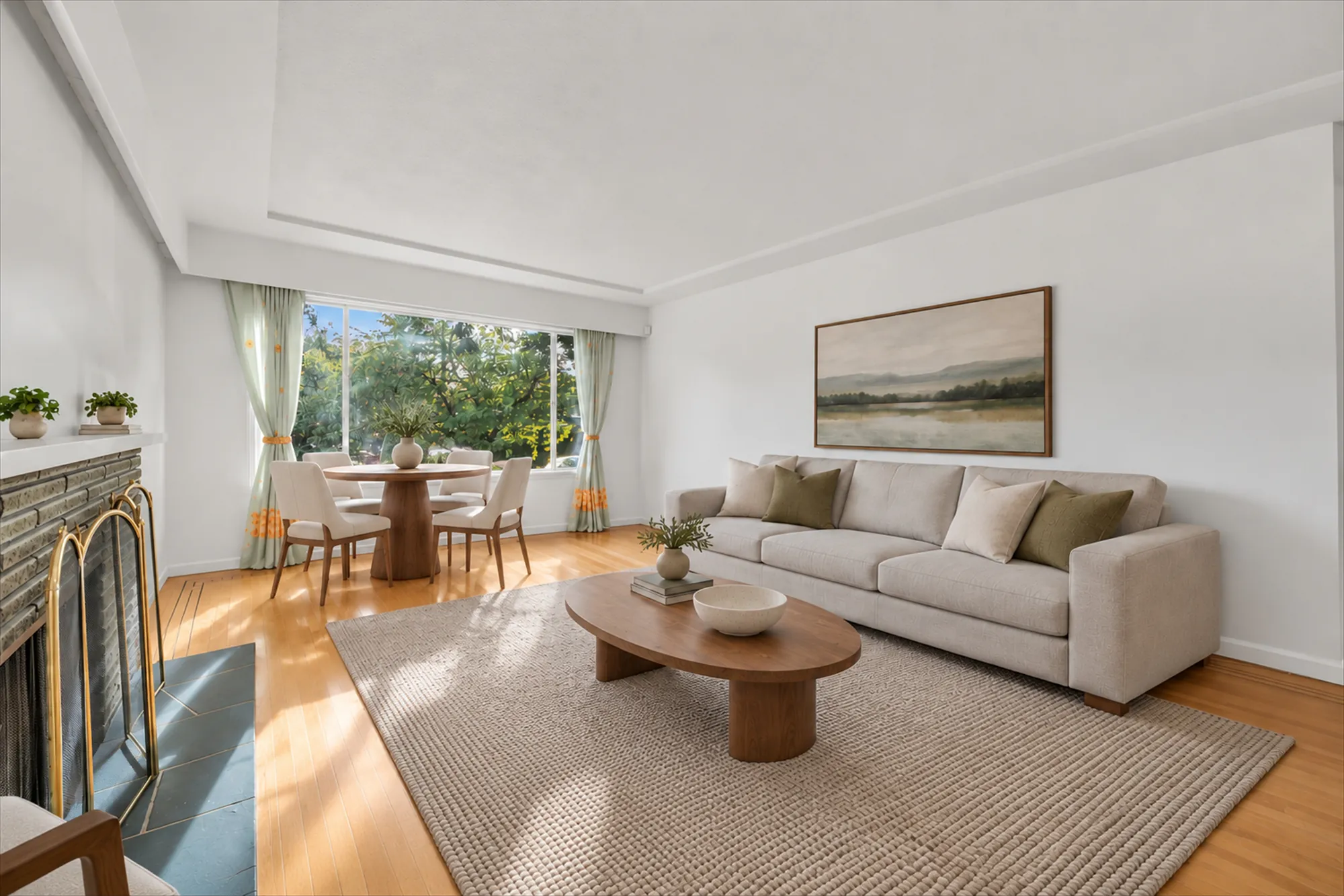 The living and dining room, virtually staged: a beige sofa and an oval coffee table on a rug, a round dining table with four chairs by a wide window, a landscape print, the fireplace with a stone surround and a brass screen at the left