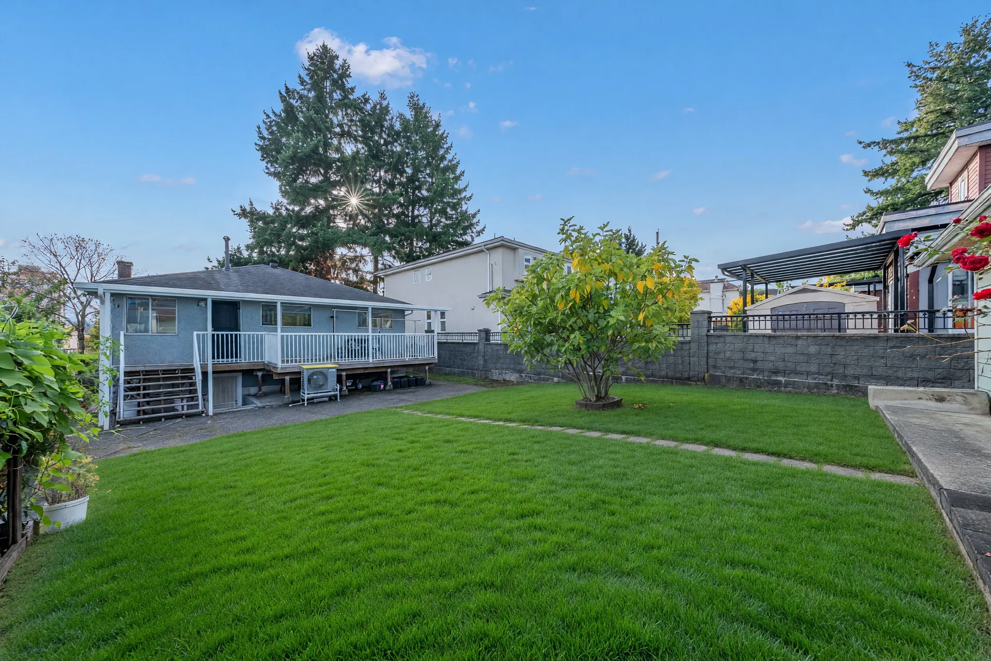 From the garage end of the lot: the lawn and a small tree, the back of the house with its covered deck, a block wall